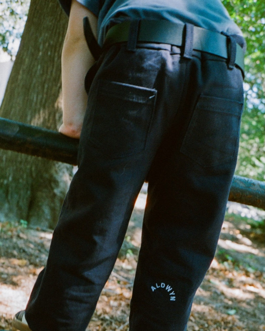 Child wearing dark pants with a brand logo, standing outdoors near a tree.