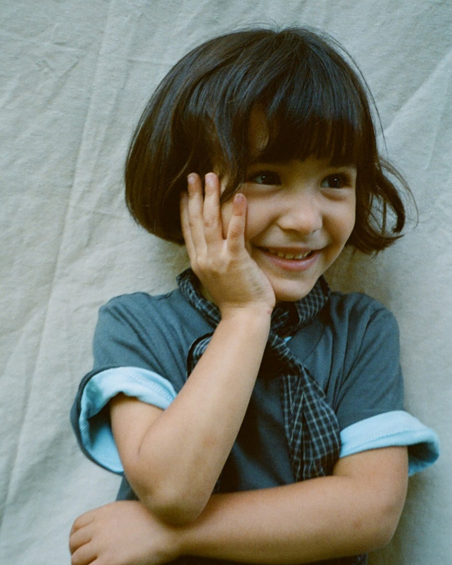 Child wearing a blue dress with white cuffs, standing against a textured light background