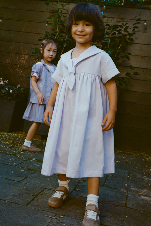 Young girl wearing a blue dress standing against a plain wall.