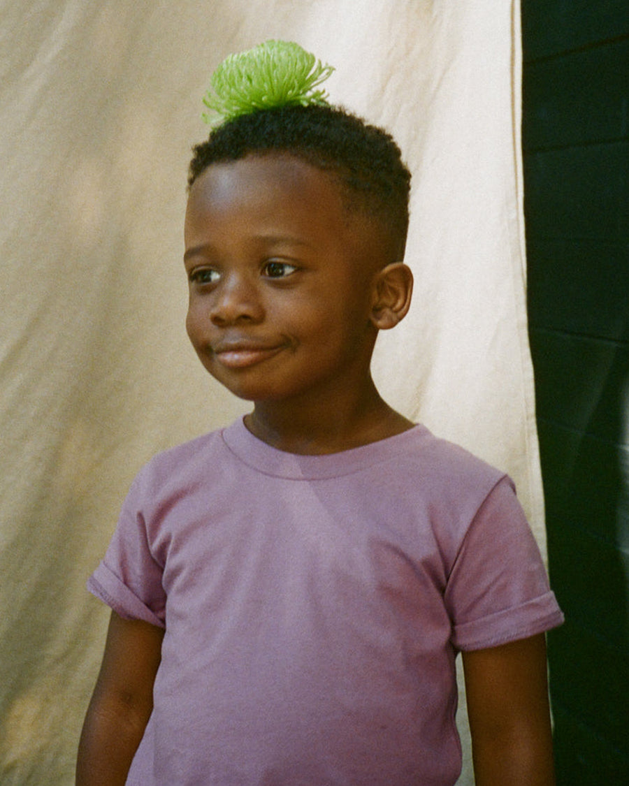 Child wearing a purple shirt with a green hair accessory against a neutral background