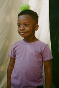 Child wearing a purple shirt with a green hair accessory against a neutral background