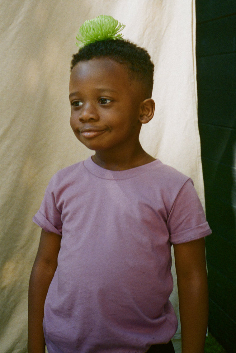 Child wearing a purple shirt with a green hair accessory against a neutral background